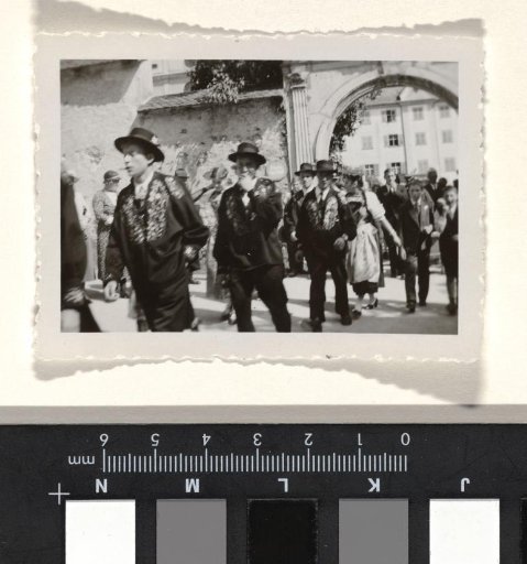 Procession of unknown men and women in dark clothing with dark hats and embroidered collars, walking under an archway