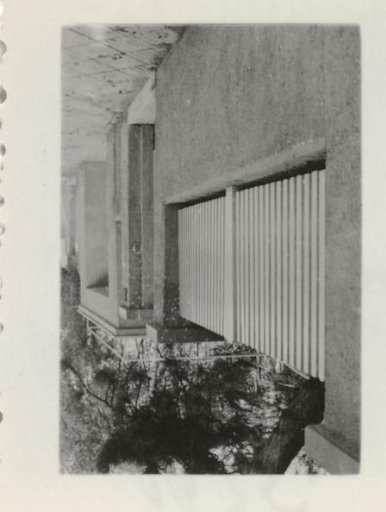 Oblique view of a stone wall with a fence on it, with trees in the background and a sidewalk in the foreground