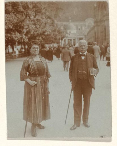 Josefine Moos-Levi and her husband Hermann Moos, both holding walking sticks, and Hermann holding a glass in his hand, standing on a street with people, buildings, and trees