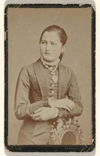Mathilde Wachenheimer-Wertheimer as a young woman in a tailored jacket with her arms folded above an embroidered chair, wearing a necklace