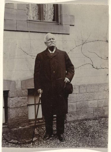 Older man with a moustache, holding a walking stick and hat in his hands, standing before a building with vegetation and two windows at knee height