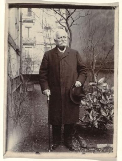 Older man with a moustache, holding a walking stick and hat in his hands, standing before a building with vegetation beside him