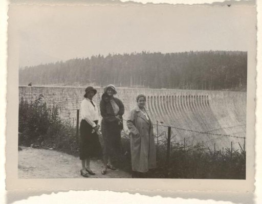 Else Wachenheimer-Moos and two unknown women wearing hats before a gate at a large concrete structure with trees in the background