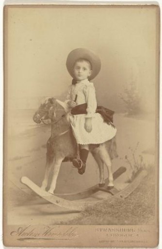 Young Eugen Wachenheimer, dressed in a dress and dark stockings, seated on a rocking horse, with a hat on his head