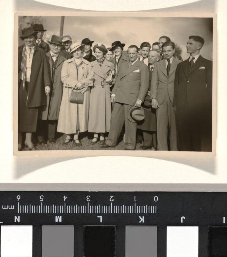 Willy Moos in a light dark suit and with a hat in his hand with 16 unknown men and women, standing outdoors with part of two trees in the background