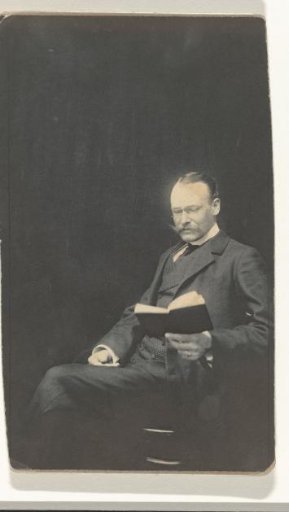 Julius Moos reading a book, seated in a chair, with a book in one hand and the other hand in his lap