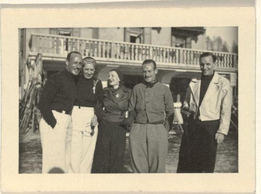 Five unknown people, with a building in the background with a balustrade and a collection of skis leaning against the building on the left