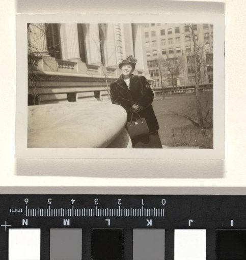 Unknown Woman, Possibly the Former Cook or Housekeeper of the Wachenheimer Family, with a Hat Adorned with Flowers, a Fur Cape and a Bag in Her Hands, Leaning Against a Large Object, with a Building with Pillars, a Tall Building and Bare Trees in the Background