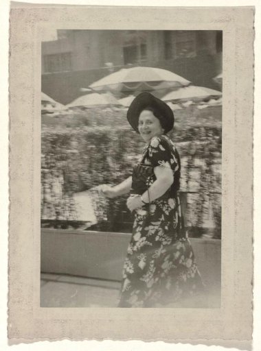 Aunt Rosel in a Dress with a Decorated Motif and a Hat on Her Head, Walking in Front of a Hedge with Parasols