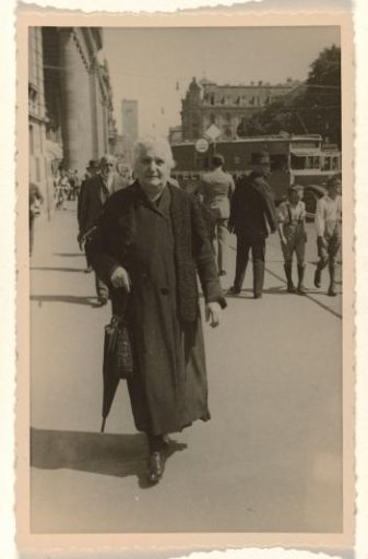 Aunt Lenchen in dark clothing with an umbrella in her hand and a shawl draped over her shoulders, walking down a street with people, buildings, and vehicles in the background