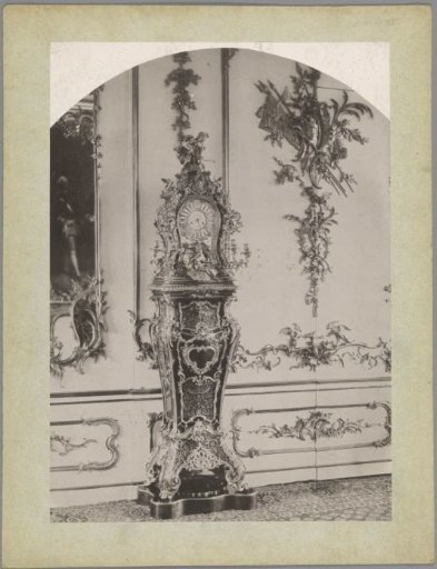 Standing clock decorated with plant motifs, a putto and a goddess, against a wall decorated with plant motifs and a family coat of arms