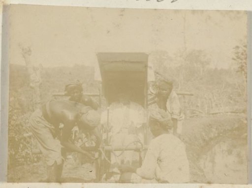 Man in a high hat in a sedan chair carried by four local men