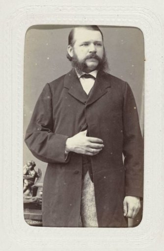 Studio portrait of a standing man with beard and mustache at a table, wearing a long coat