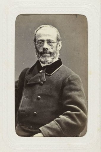 Studio portrait of a man with glasses, mustache, and beard in a long woolen coat with a velvet collar