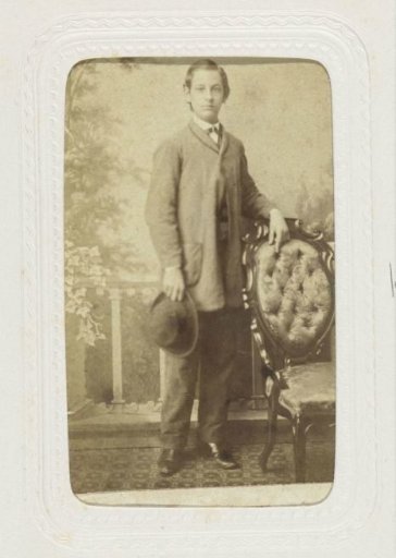 Studio portrait of a standing man with his right hand in the breast of his coat and holding a book in his other hand