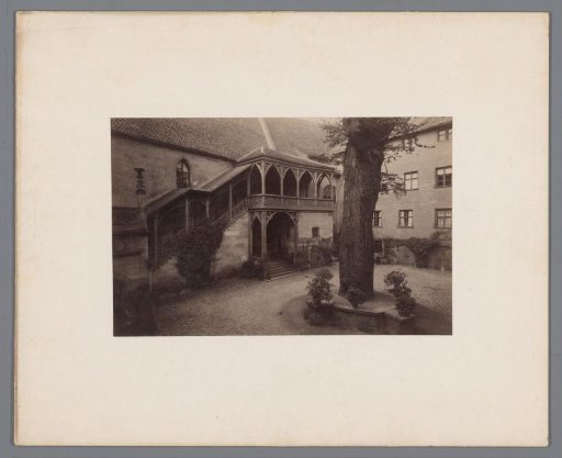 View of a courtyard with a tall tree, houses, and a cloister with pointed arches and a veranda