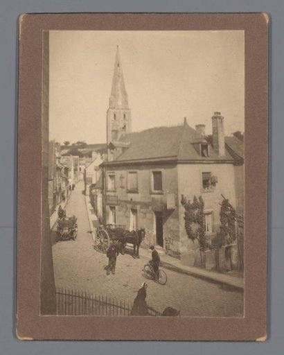 View of a Belgian (?) town or village, with a church, houses, and on the street a car, next to a carriage and a cyclist
