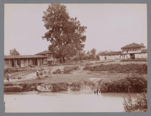 Photograph of two large houses in a grassy field with water [pond] in the foreground, Iran