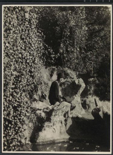 Portrait of G.A. Urtebise in a dark dress with a bowler hat sitting on a rock by a pond