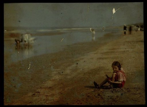 Girl (dressed in red, seated) playing on the North Sea beach, with various figures in the background