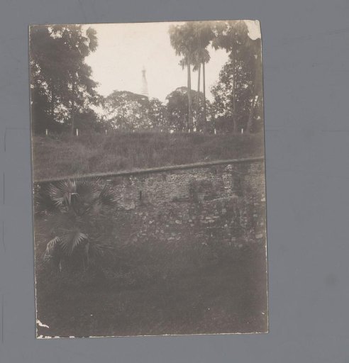View of a stone wall against a hill with the top of a Buddhist shrine behind it, Myanmar