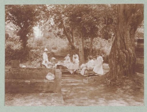 A set table in the tropics with Europeans dressed in white seated at it.