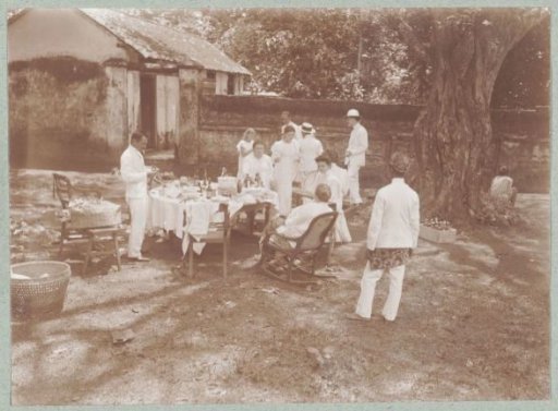 Europeans dressed in white relaxing at an outdoor table while an Indonesian servant watches from a distance.