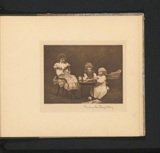 Two girls at a table in a garden with a toy tea set, the eldest reading to a doll and the younger girl
