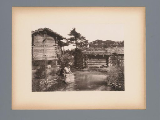 View of Log Cabins and a Woman Washing Clothes in the Water