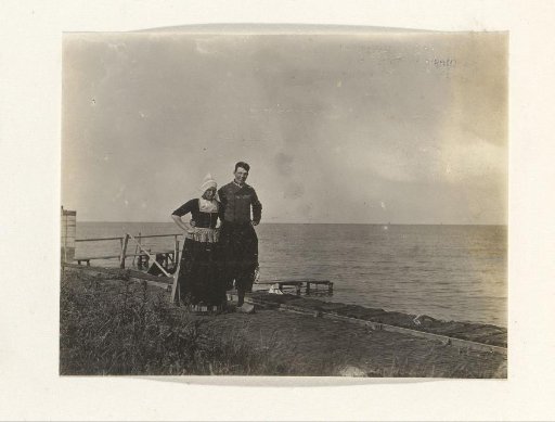 Two men in traditional dress (one in women's clothing) by the water