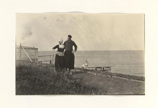 Two men in traditional dress (one in women's clothing) by the water