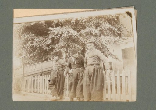 Three men in traditional dress, standing before a fence