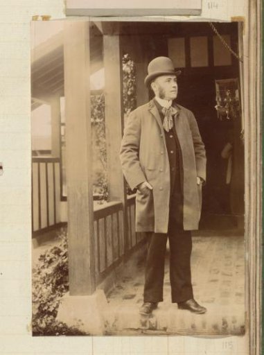 Portrait of a man with a round hat and coat, standing by the veranda of a house