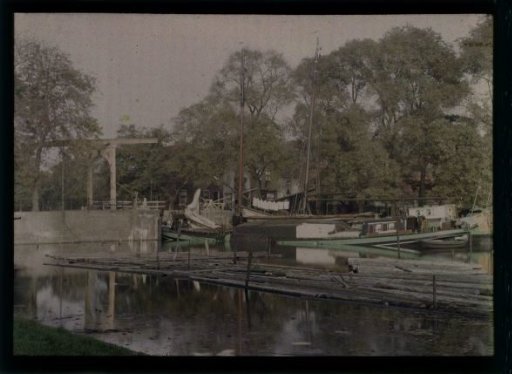 View of a canal with floating timber in the foreground, a ship, a drawbridge (left), hanging laundry, and the houses and/or farmhouses of a village, North Holland (?)