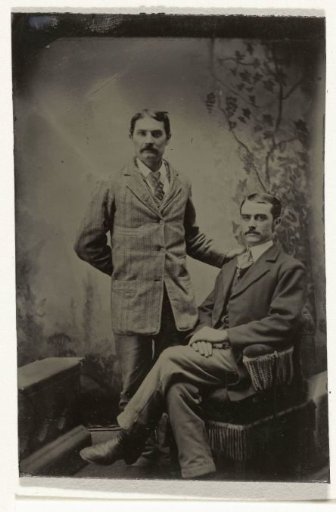 Portrait of two men, one standing and one sitting, in front of a painted backdrop (leaves on a tree branch)