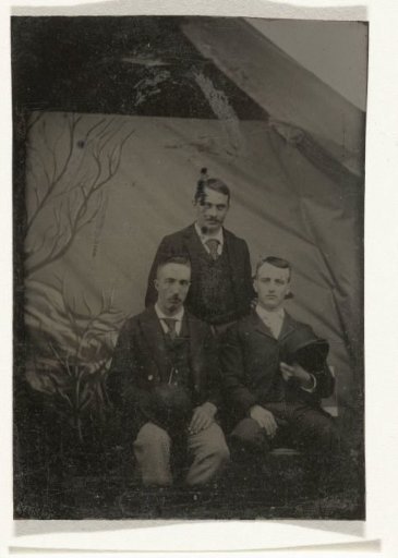 Portrait of three men, standing and sitting in front of a painted backdrop (tree branches)