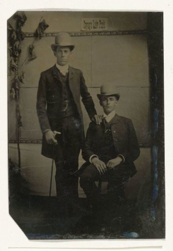 Portrait of two men, one standing and one sitting in a studio, with a paper on the back wall that reads Cayuga Lake Park (= Cayuga Lake State Park), NY, United States