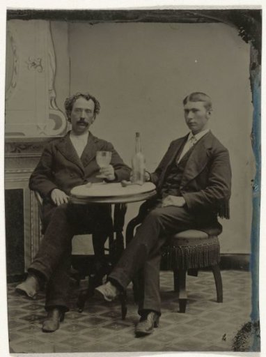 Portrait of Two Men Sitting at a Table in a Studio, with a Bottle and a Glass, in Front of a Faux Salon Decor