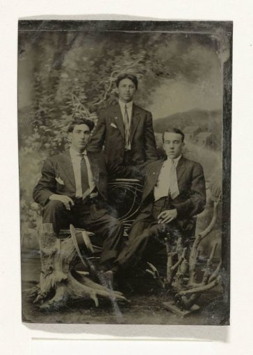 Portrait of three men, two of whom hold cigars, standing and seated before a painted backdrop