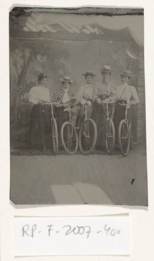 Group portrait of five women, standing with bicycles, in front of a painted backdrop depicting a forest landscape, with part of the photographer's tent visible above