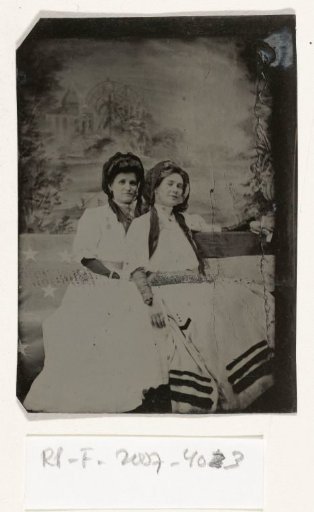 Portrait of two women sitting on a bench in front of a painted backdrop (chapel or church (?) by a lake), with part of the American flag on the left