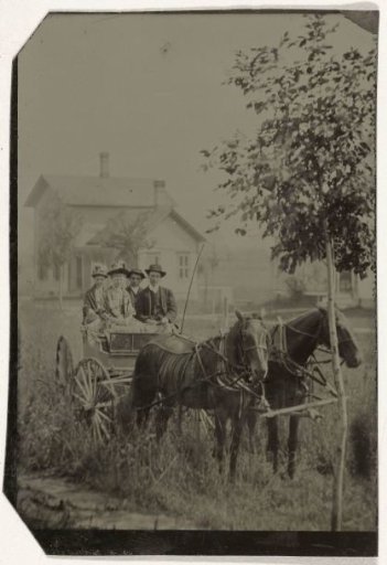 Two men and two women in a carriage with two horses, with a wooden house in the background