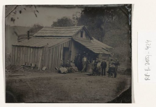 View of a wooden house with ten people, one of whom is being cut or shaved