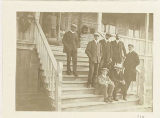 Portrait of a company of Dolph Kessler's traveling companions on the steps in front of a wooden house
