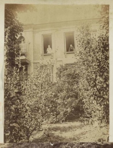 Facade of a house with three people in the windows, seen from a garden in France