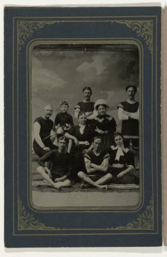 Group portrait of five men, three women, and a girl in bathing attire, before a painted backdrop with a cloudy sky
