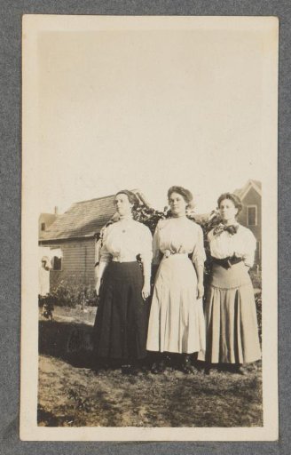Three young women near some houses, laundry hanging on the left