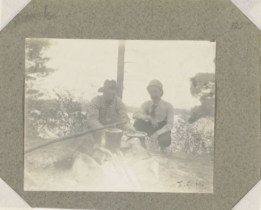 Two men, crouching, cooking over a campfire