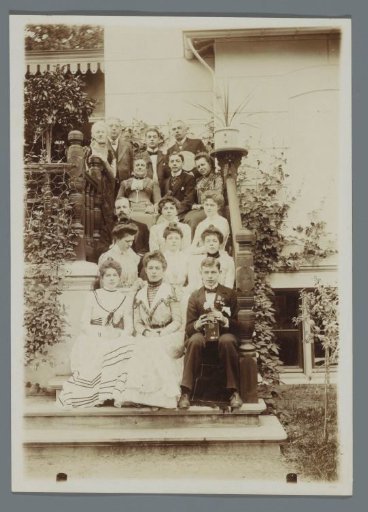 Group portrait of the Geertsema family with Karel van den Berg, seated on the steps behind the house; young man with camera in the front right