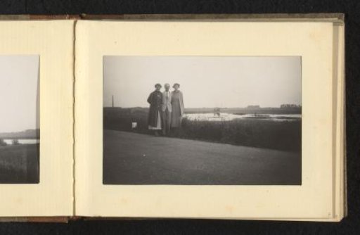 Man with Two Women on Either Side, on a Paved Road Next to a Lake or River